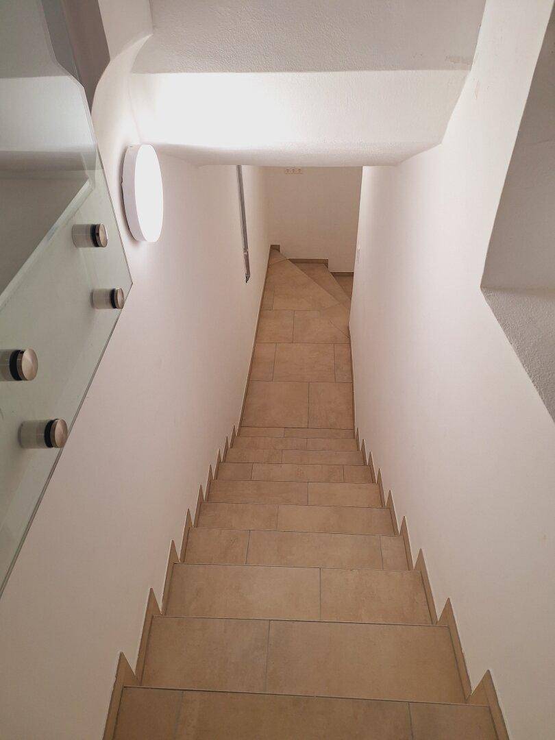 Modernes Treppenhaus mit Fliesenstufen und einem eleganten Glasgeländer, das nach unten führt.