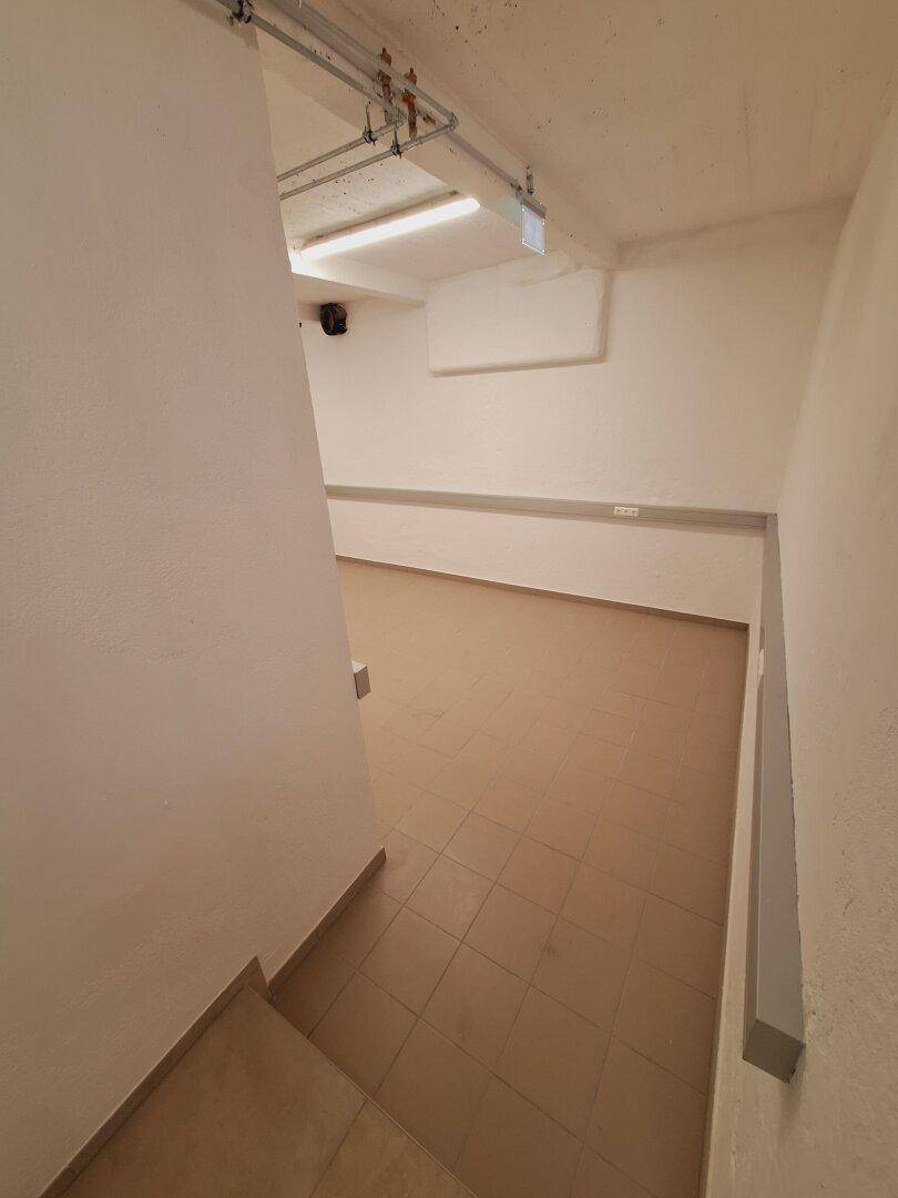 Kellerraum mit Fliesenboden, weiß gestrichenen Wänden und sichtbaren Leitungen an der Decke.