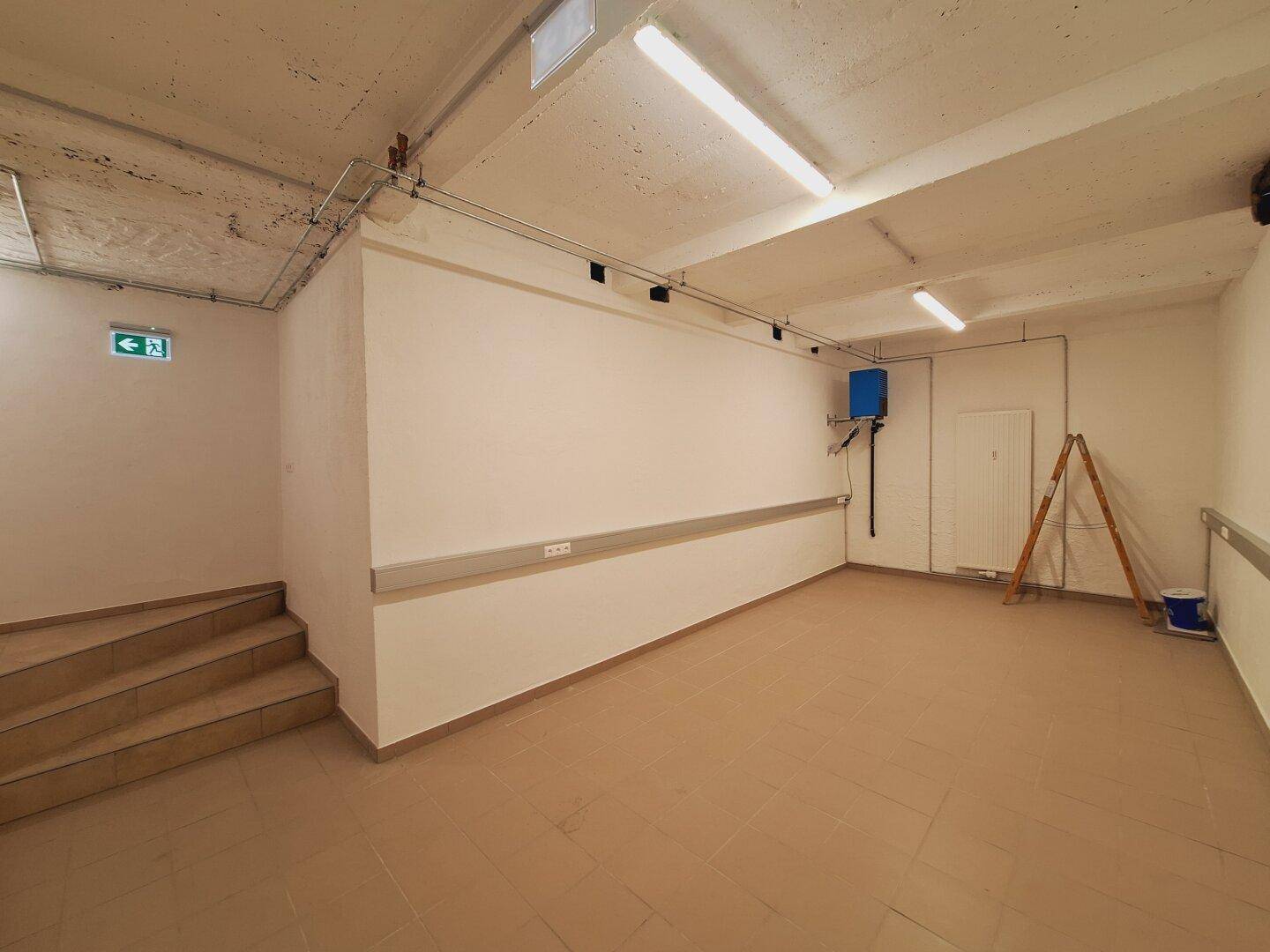 Kellerraum mit Fliesenboden, Treppenzugang und sichtbaren Leitungen an der Decke und Wänden.