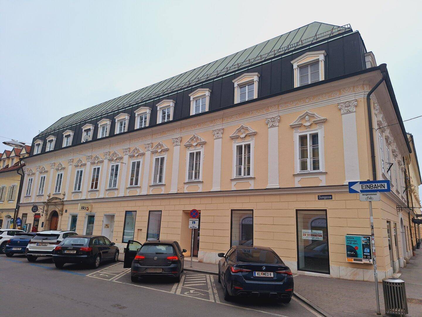Historisches Gebäude mit Geschäften im Erdgeschoss und parkenden Autos auf der Straße an einem bewölkten Tag.
