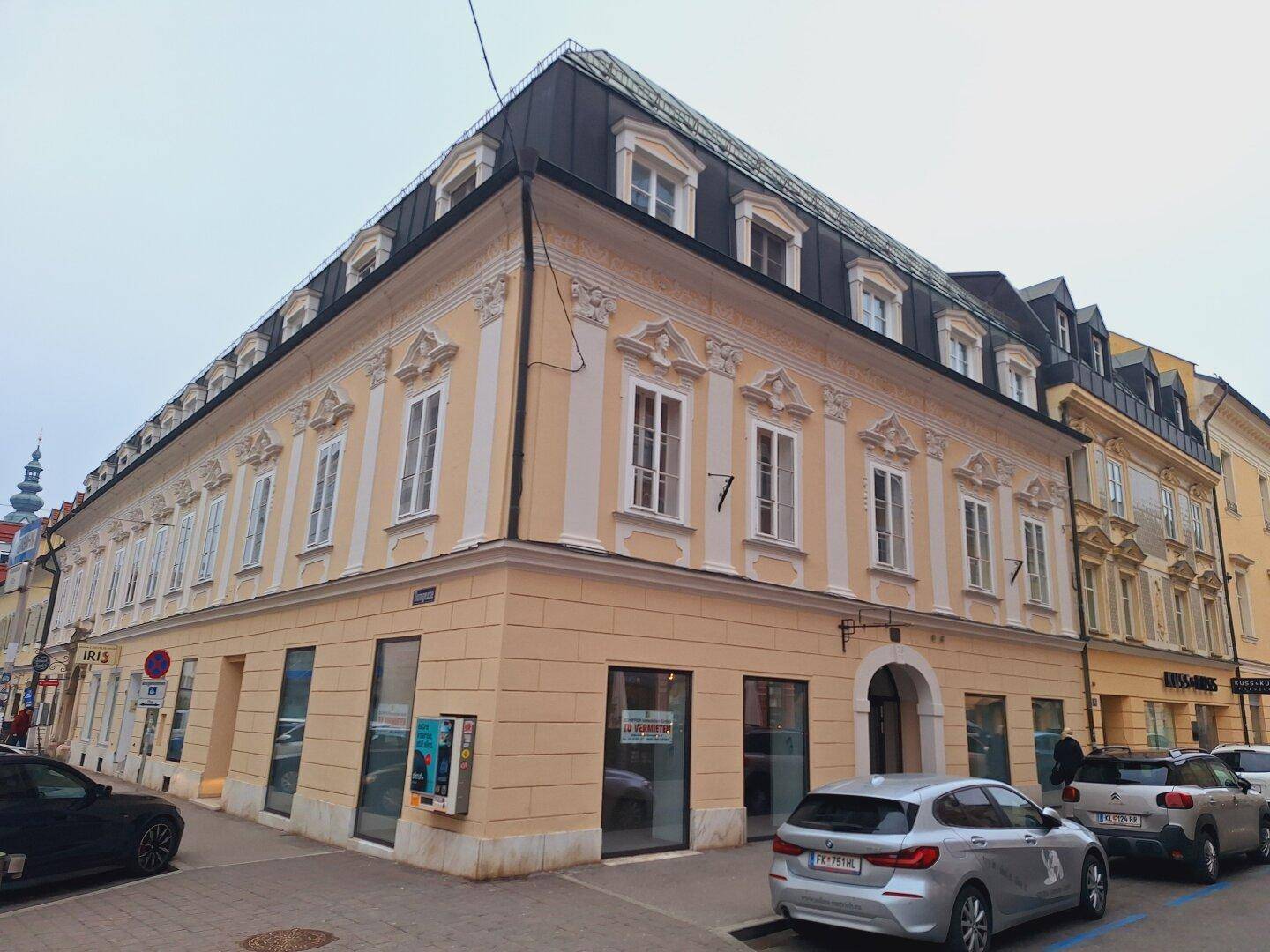 Historisches Eckgebäude mit klassischer Fassade, großen Fenstern und Geschäften im Erdgeschoss an einer belebten Straße.