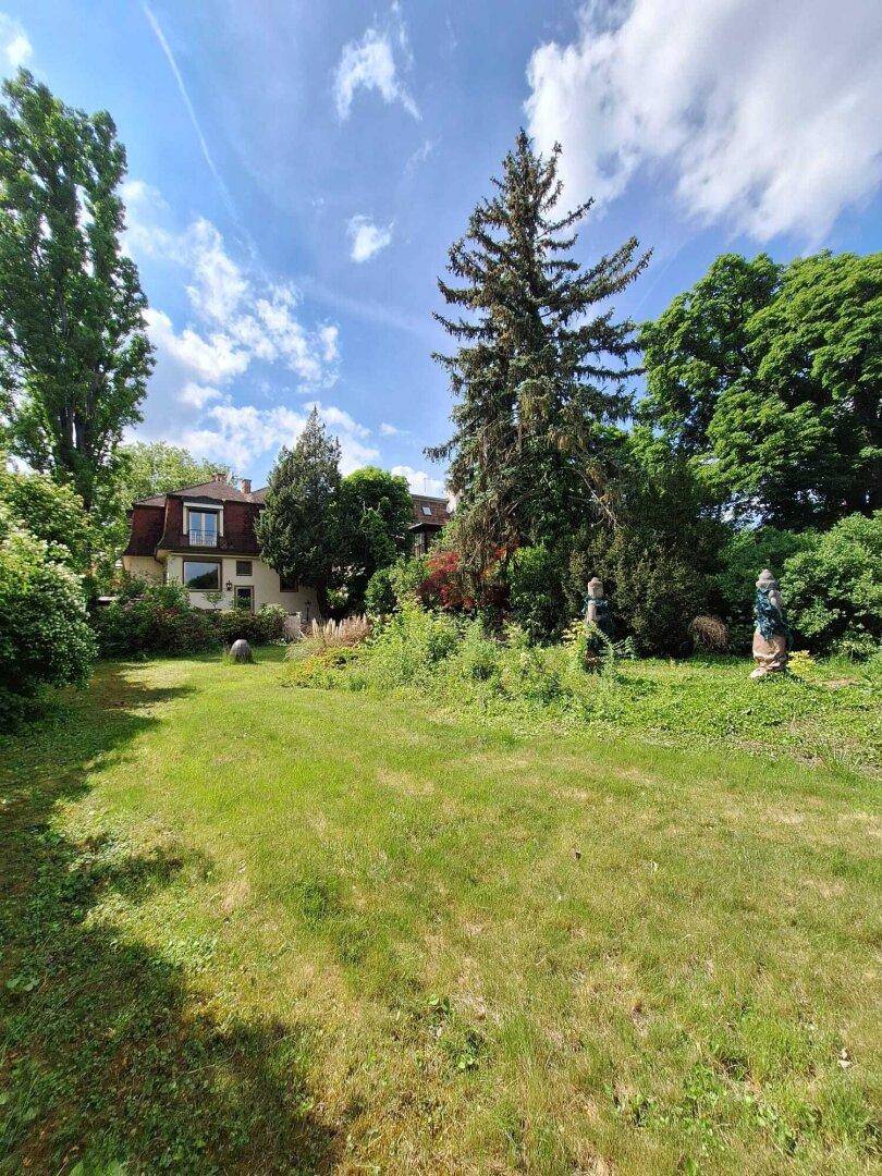 Weitläufiger Garten mit Rasenfläche, altem Baumbestand und dekorativen Elementen unter blauem Himmel.