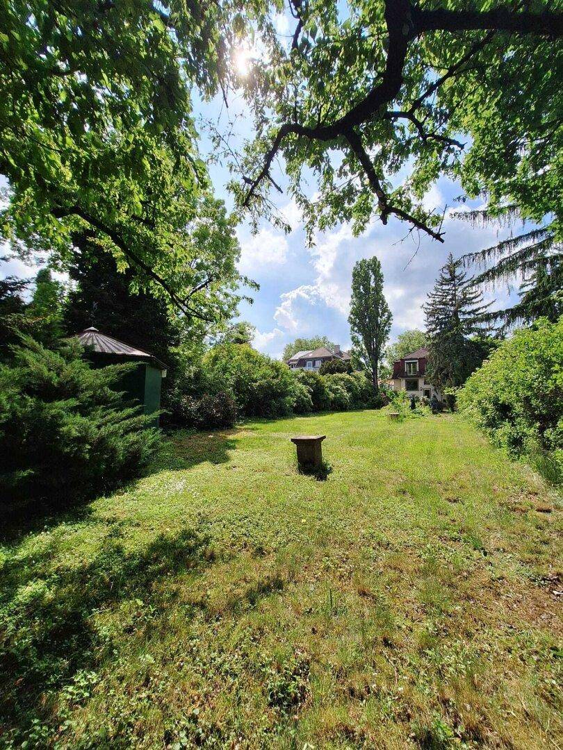 Weitläufiger Gartenbereich mit Rasen, altem Baumbestand und einem kleinen Gartenhaus im Hintergrund.