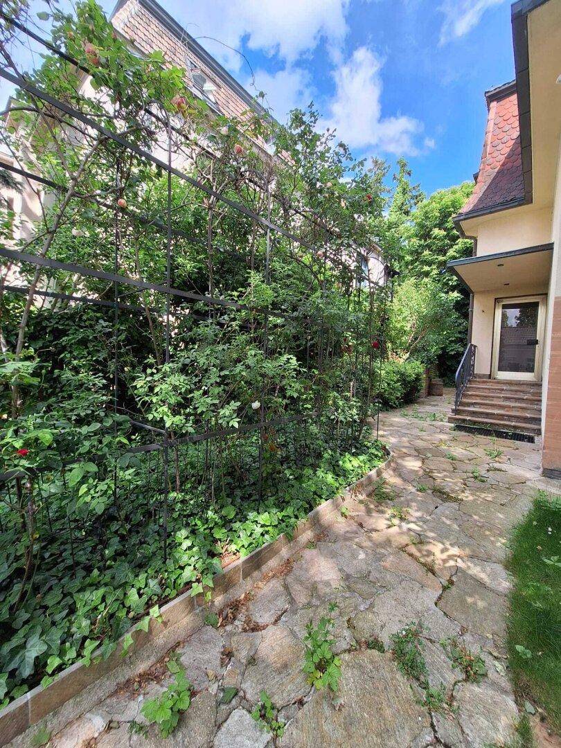 Gepflasterter Weg entlang des Hauses mit einer bewachsenen Pergola und üppigem Grün.