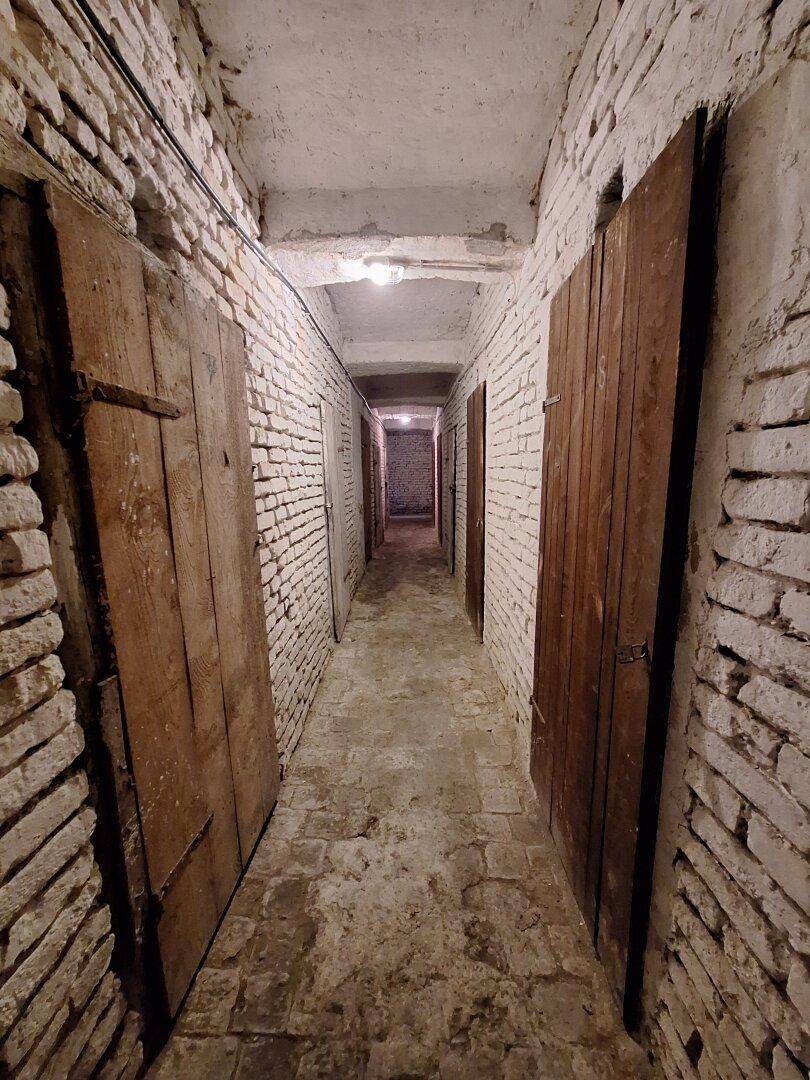 Langer Kellergang mit unverputzten Ziegelwänden und Holztüren zu den Abteilen.