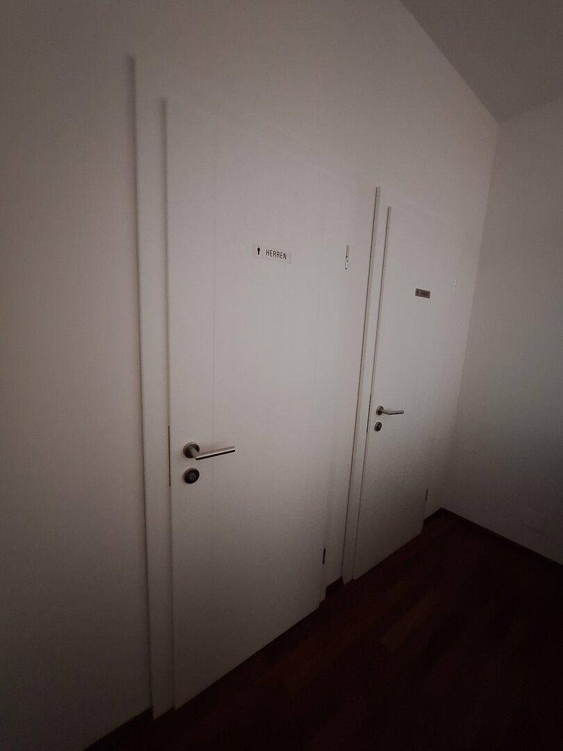 Zwei weiße Türen, eine davon mit der Aufschrift "HERREN", in einem schlichten Innenbereich.