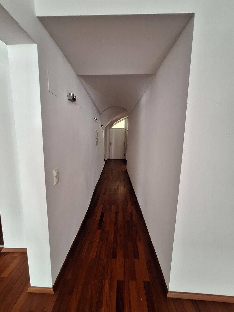 Langer, schmaler Flur mit weißen Wänden und dunklem Holzparkett, der zu einem weiteren Bereich führt.