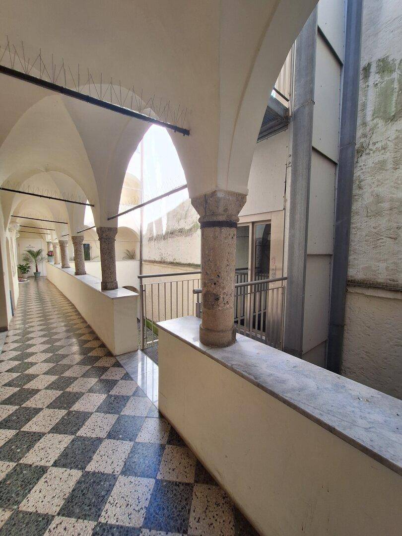 Lange, gewölbte Loggia mit einer Reihe von Steinpfeilern und einem auffälligen Schachbrettmuster-Fliesenboden.
