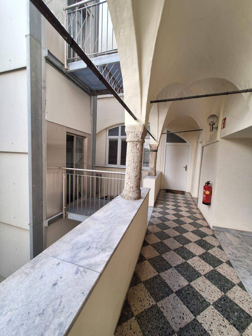 Historische Loggia mit Steinpfeilern, Gewölbedecke und einem schwarz-weiß gefliesten Boden, angrenzend an eine Außentreppe.