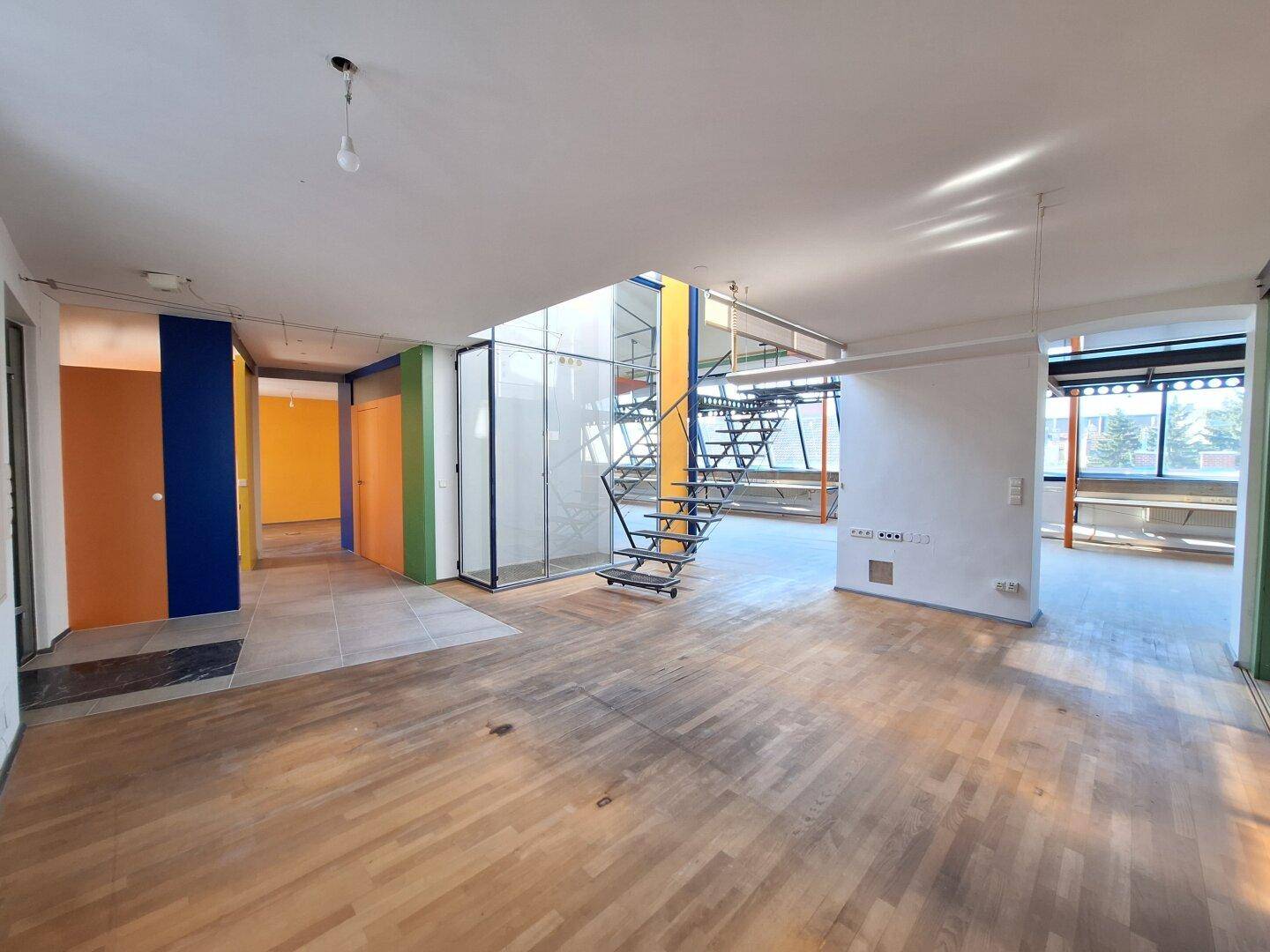 Geräumiger offener Bürobereich mit Holzboden, bunten Wänden und einer modernen Metalltreppe.