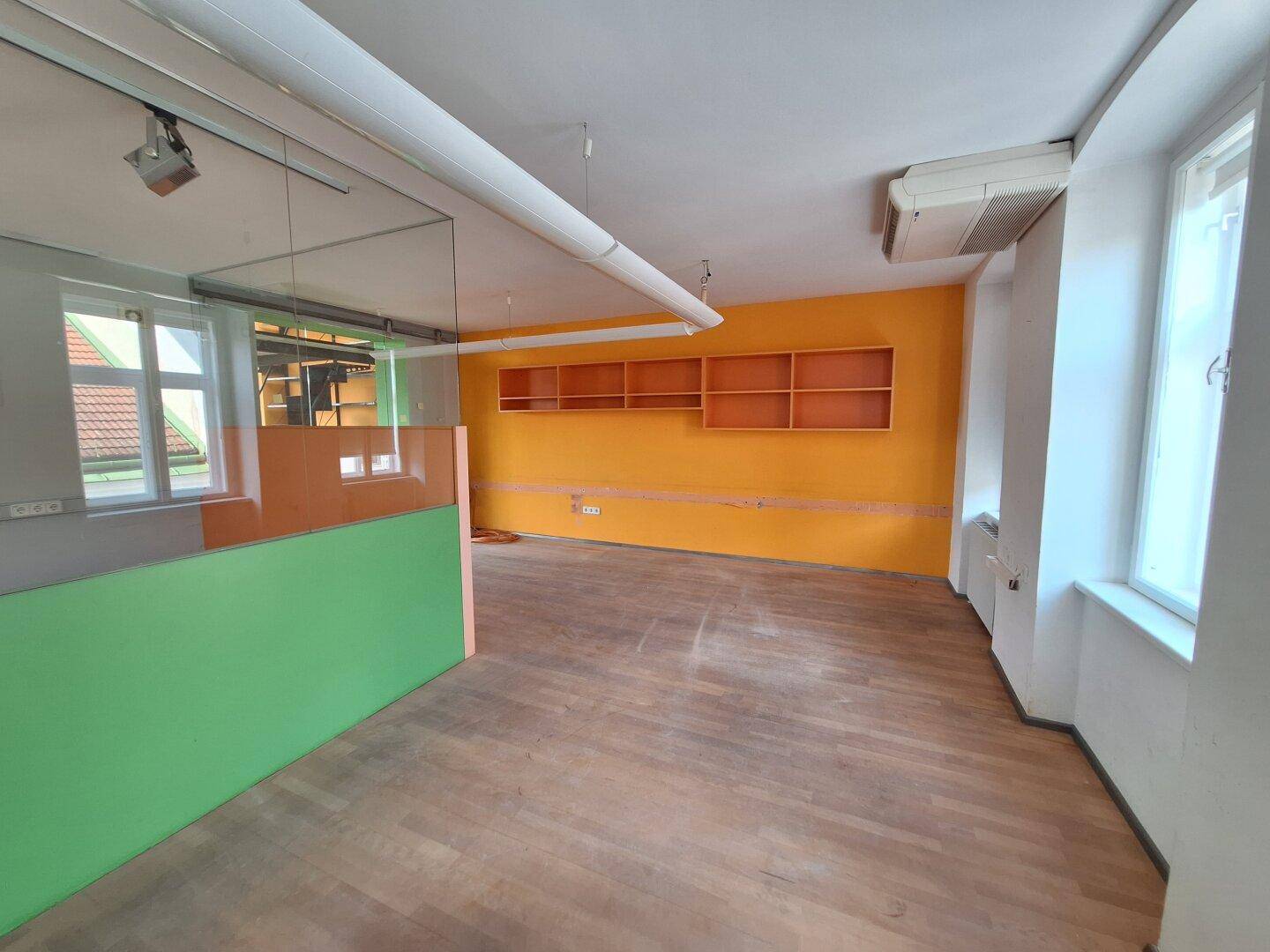 Büroraum mit Holzboden, orangefarbener Akzentwand und integrierten Regalen, gut beleuchtet.