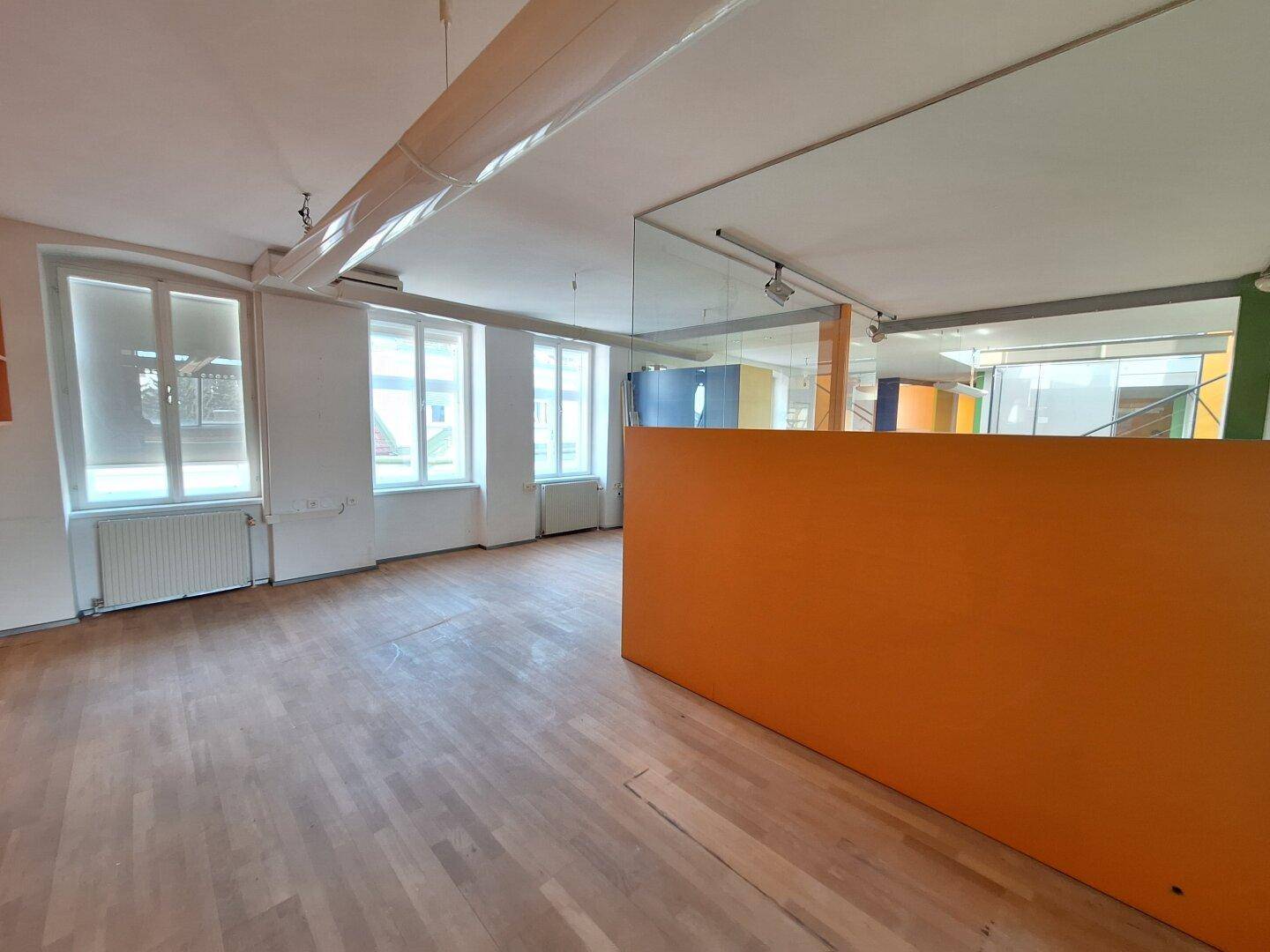 Heller Büroraum mit Holzboden, großen Fenstern und einer orangefarbenen Akzentwand.