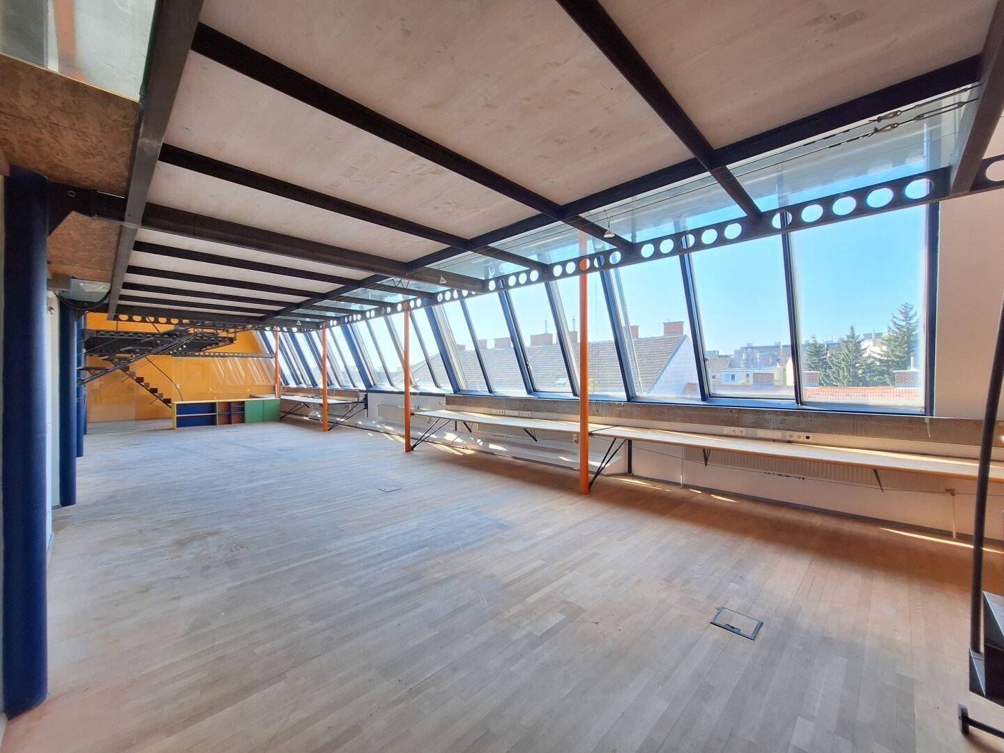 Weitläufiger Büroraum mit Holzboden, schrägen Glasfenstern und Blick auf die Dächer der Umgebung.