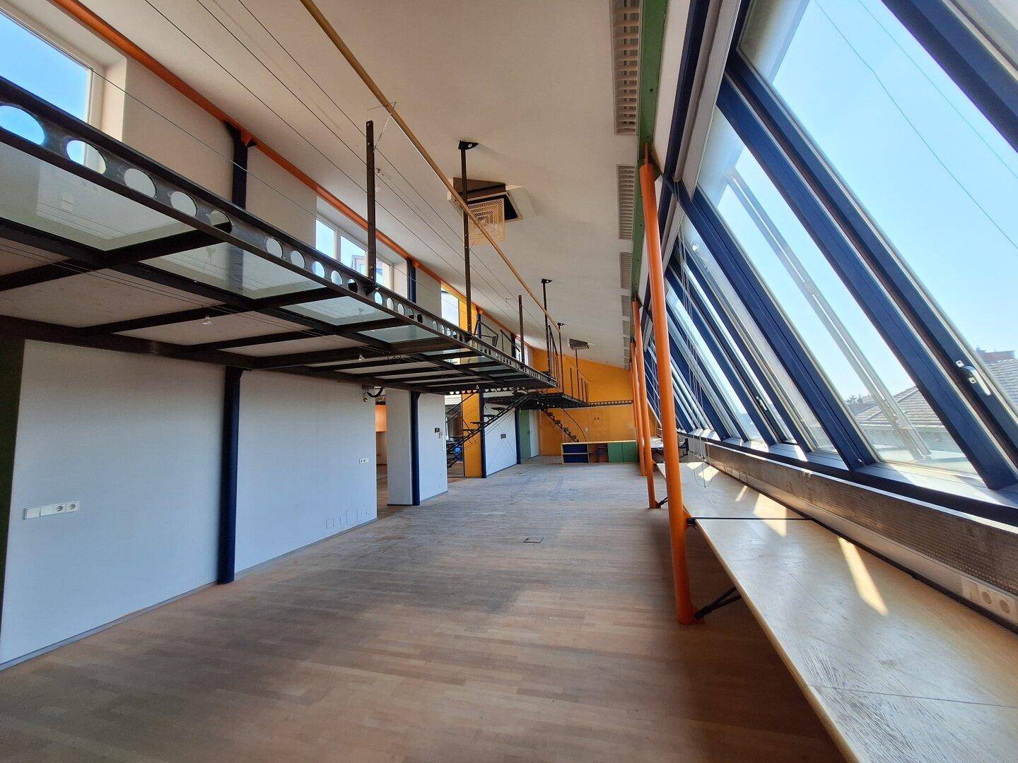 Langer Bürobereich mit Holzboden, schrägen Glasfenstern und einer erhöhten Metallbrücke.
