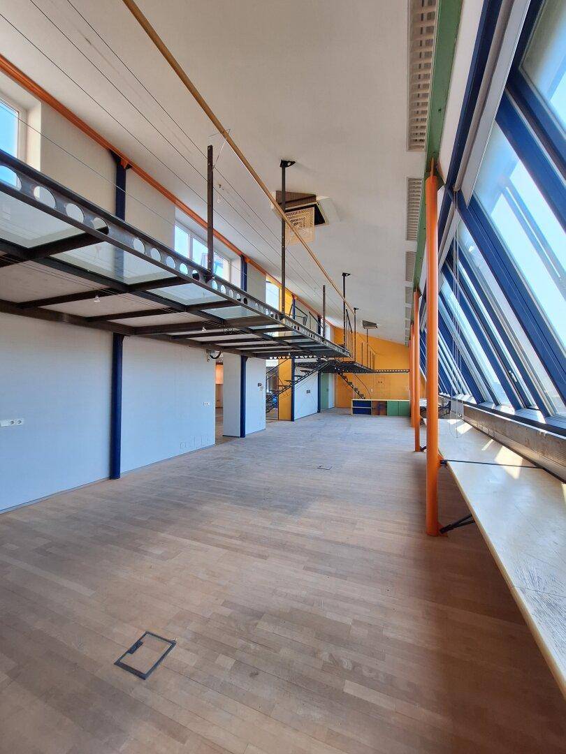 Langer Bürobereich mit Holzboden, schrägen Glasfenstern und einer erhöhten Metallbrücke.