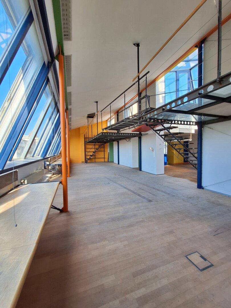 Geräumiger Bürobereich mit Holzboden, schrägen Glasfenstern und einer erhöhten Metallbrücke.