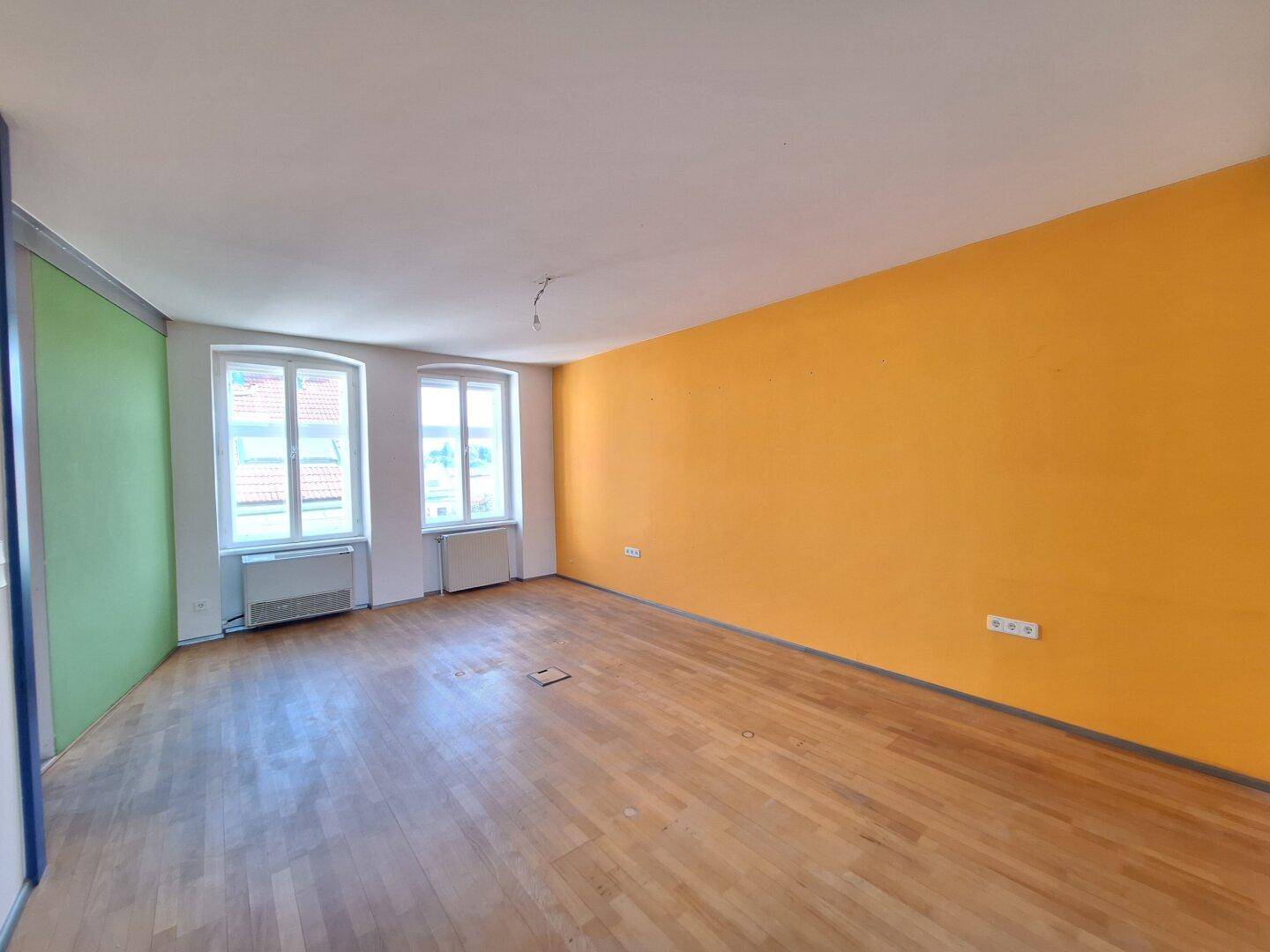 Heller Büroraum mit Holzboden, großen Fenstern und einer orangefarbenen Akzentwand.