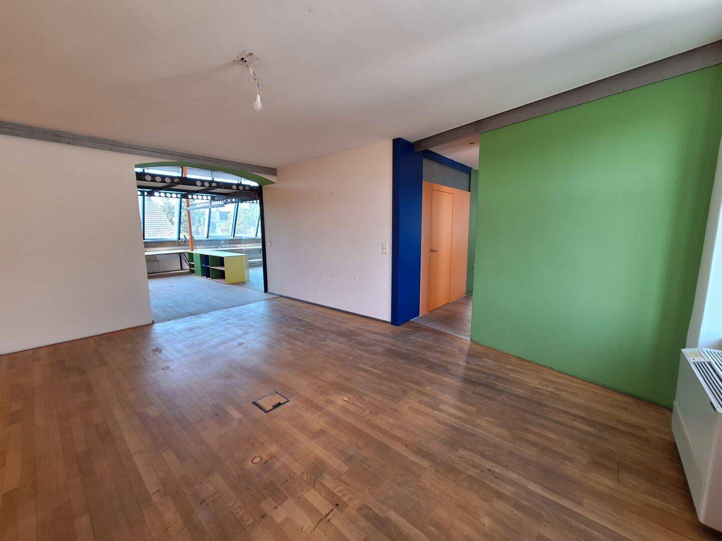 Offener Bürobereich mit Holzboden, grüner Akzentwand und Zugang zu einem weiteren Raum.