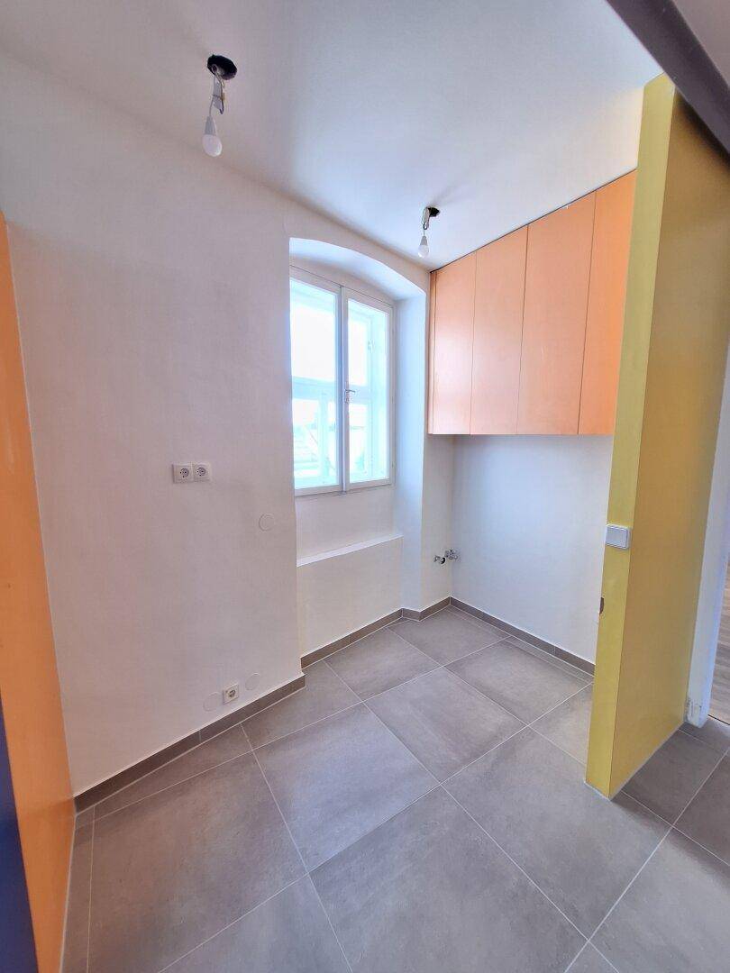 Kleiner Raum mit Fliesenboden, einem Fenster und orangefarbenen Hängeschränken.