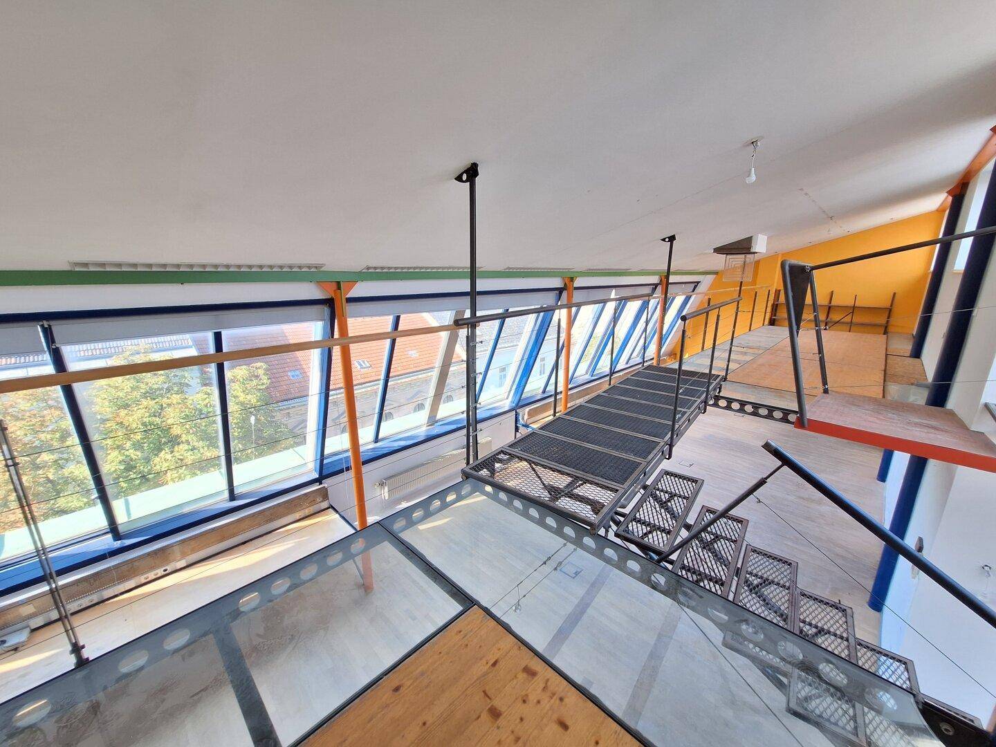 Blick von oben auf den offenen Bürobereich mit Metallbrücke, Treppe und schrägen Glasfenstern.