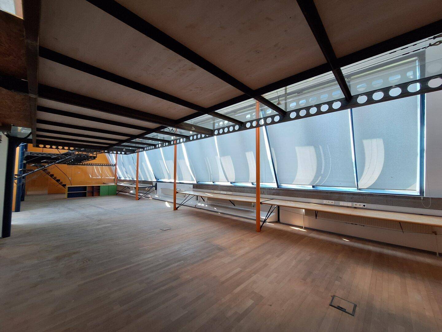 Langer Innenraum mit sichtbaren Deckenbalken, großen Fenstern mit Rollos und einer durchgehenden Fensterbank.