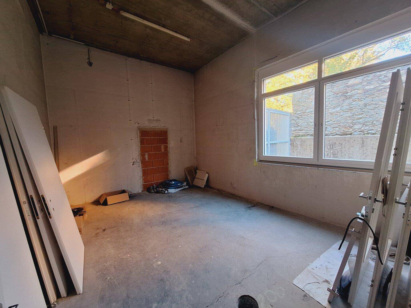 Kleinerer Gewerberaum im Rohbau mit Fenster und Blick auf eine Steinmauer.