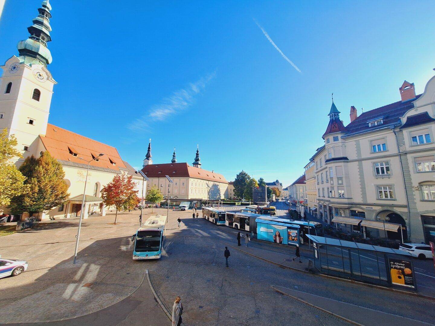 Historischer Stadtplatz mit Kirche, modernen Gebäuden und Bussen unter strahlend blauem Himmel.