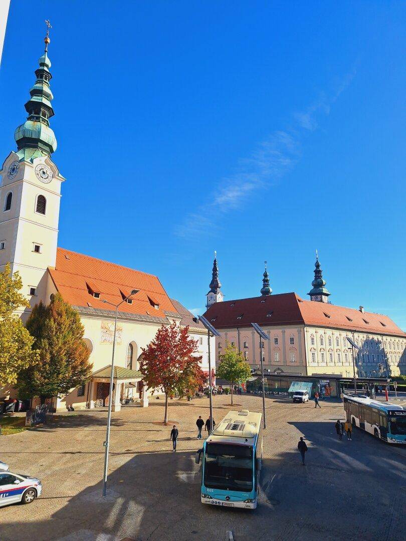 Historischer Stadtplatz mit Kirche, Gebäuden und Bussen unter strahlend blauem Himmel.