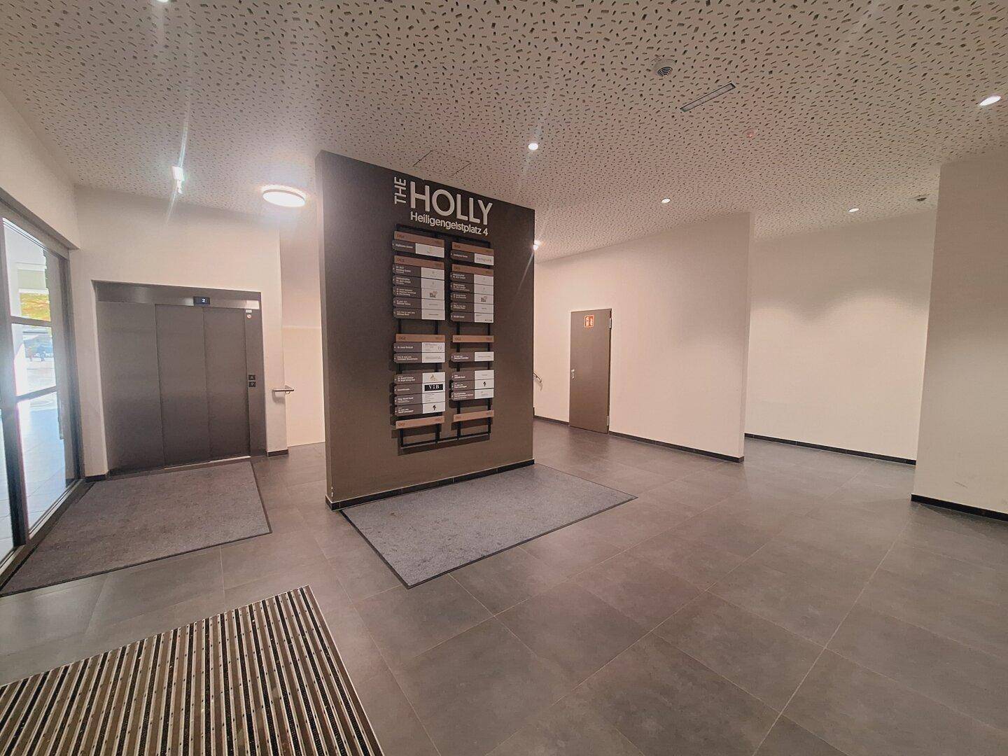 Moderner Eingangsbereich mit Aufzug, Informationswand und dunklem Fliesenboden.