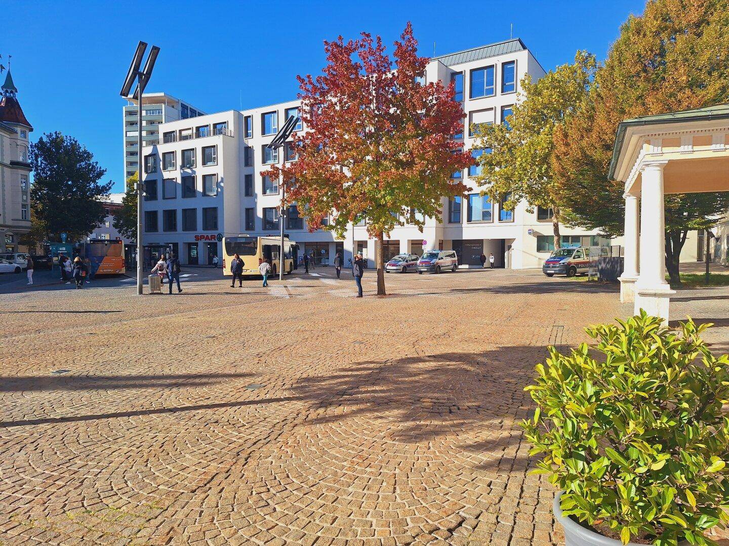 Belebtes Stadtzentrum mit modernen Gebäuden, Kopfsteinpflaster und Herbstbäumen unter klarem Himmel.