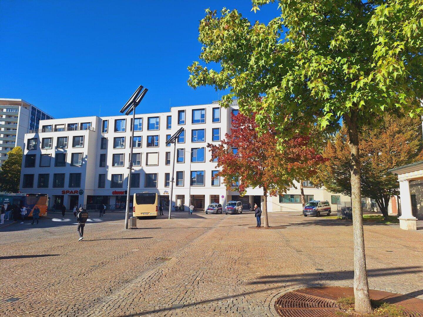 Ein belebter Stadtplatz mit modernen Gebäuden, Bäumen und einem Bus an einem sonnigen Herbsttag.