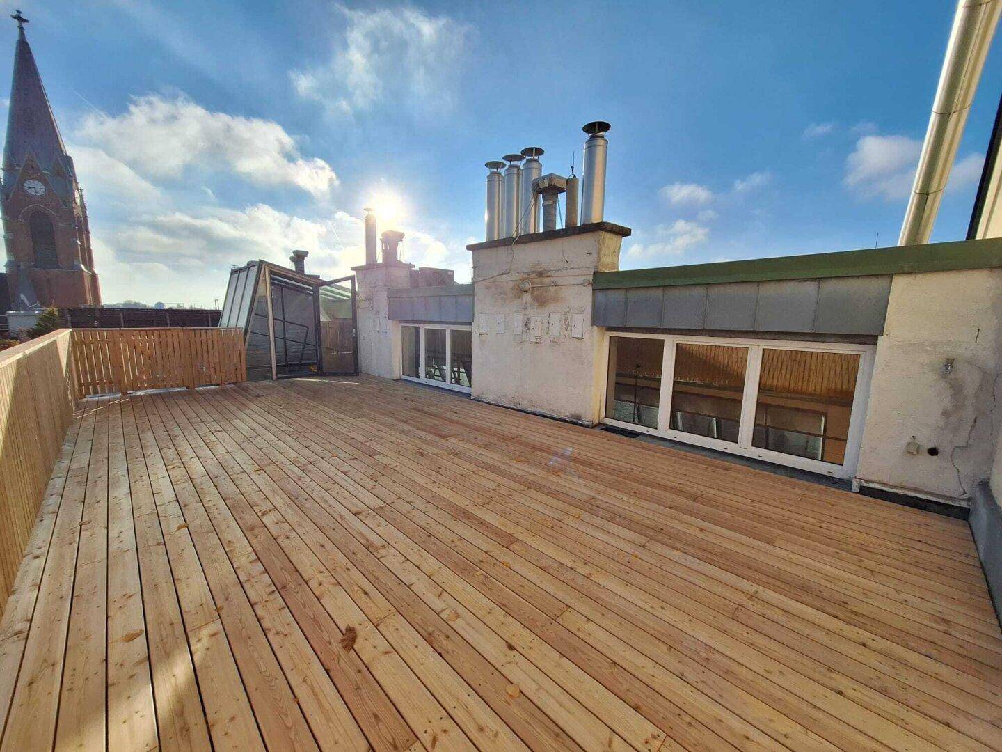 Sonnige Dachterrasse mit Holzbelag, Holzzaun und Blick auf die Dächer und einen Kirchturm.