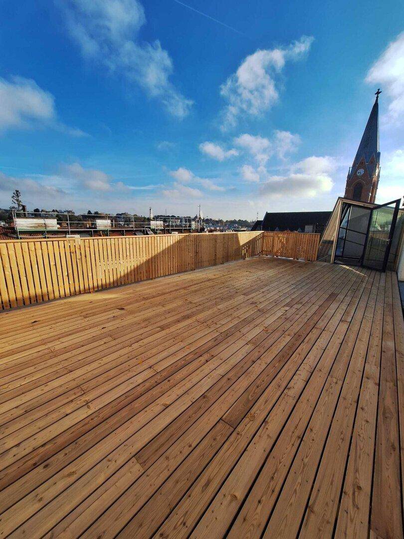 Weitläufige Dachterrasse mit neuem Holzbelag, Holzzaun und Blick auf die Stadt mit Kirchturm.