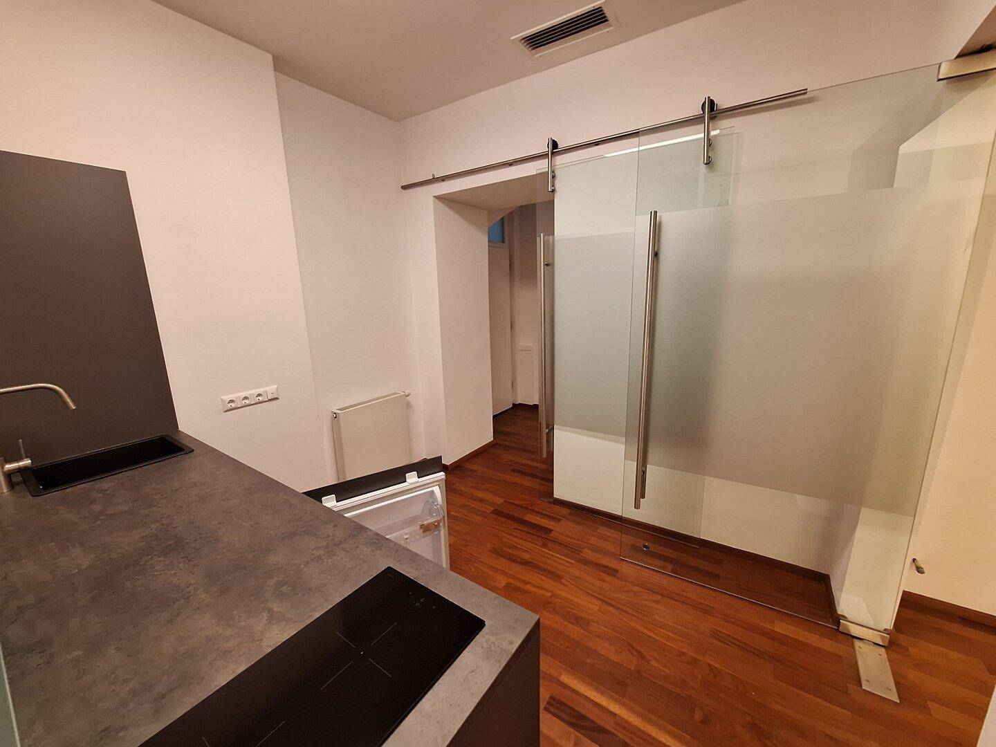 Kompakte Küchenzeile mit Induktionskochfeld, Spüle und kleinem Kühlschrank, abgetrennt durch eine Glasschiebetür.