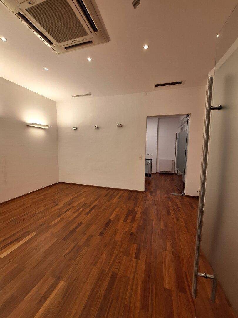 Heller Büroraum mit Holzboden und Wandleuchten, der durch eine Glastür zugänglich ist.