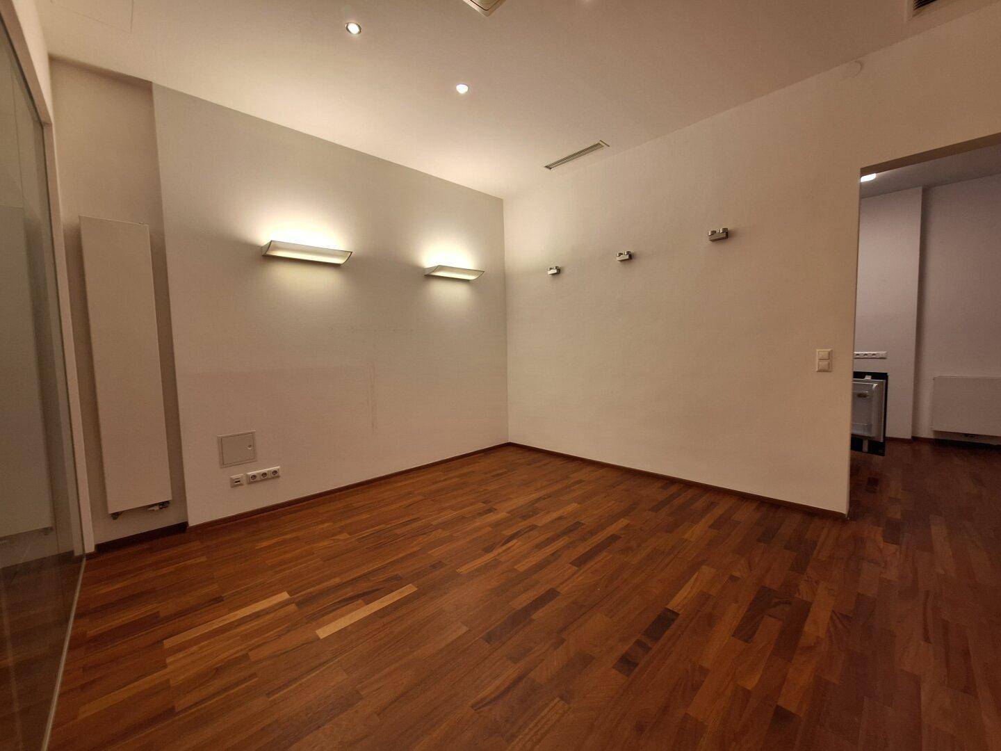Heller Büroraum mit Holzboden und Wandleuchten, der durch eine Glastür zugänglich ist.