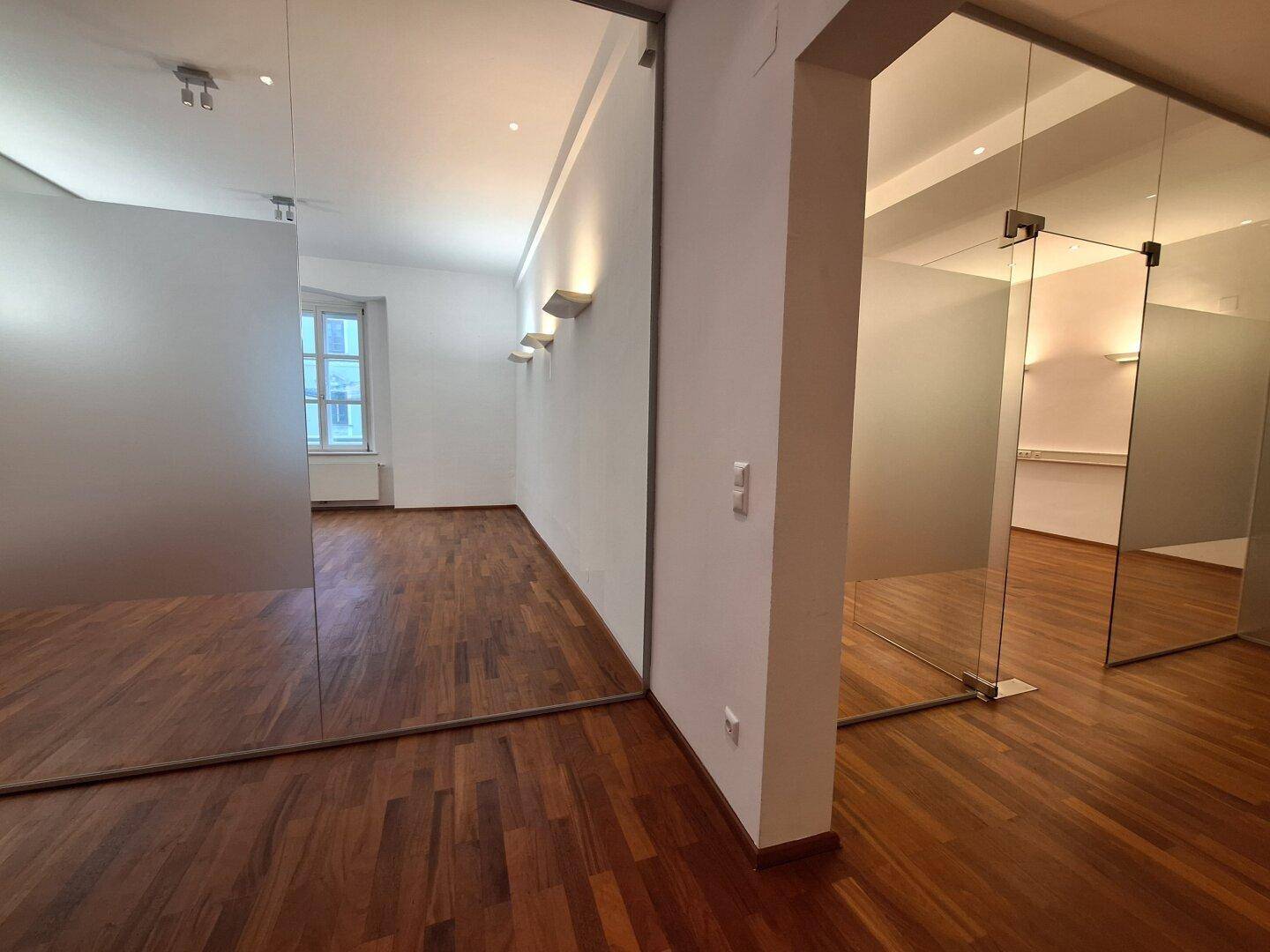 Heller Büroraum mit Holzboden und Wandleuchten, der durch eine Glastür zugänglich ist.