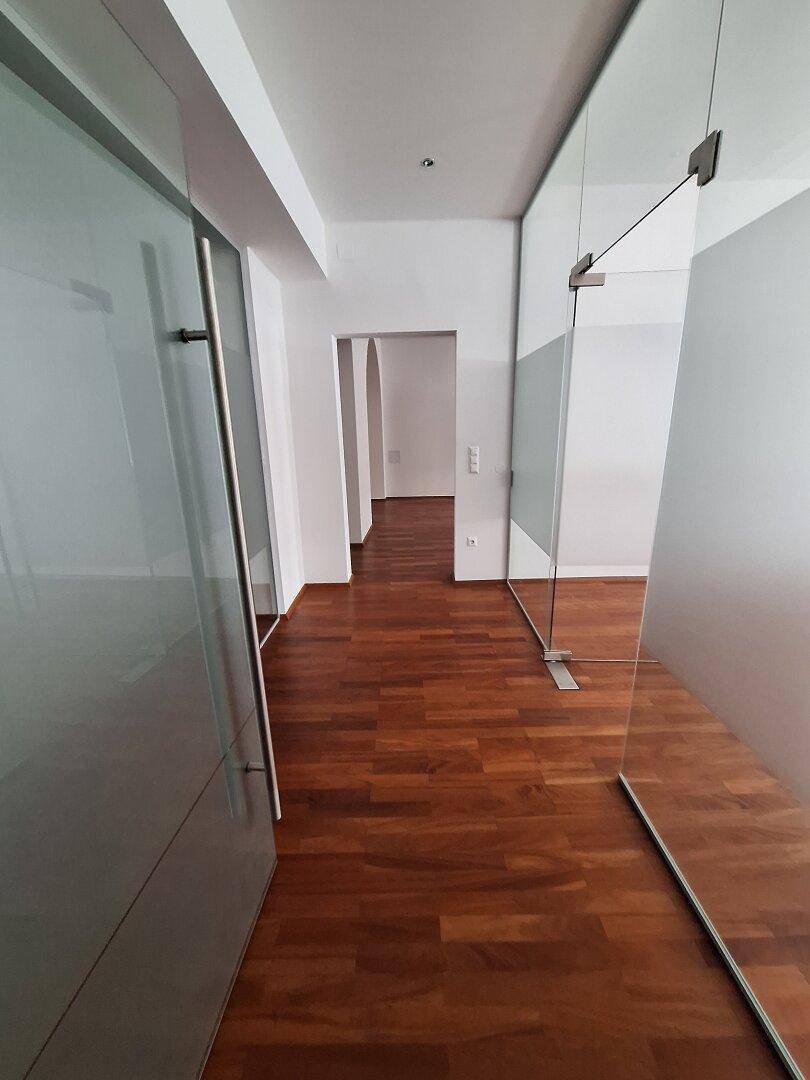 Heller Flur mit Holzboden und Glaswänden, der zu den verschiedenen Büroräumen führt.