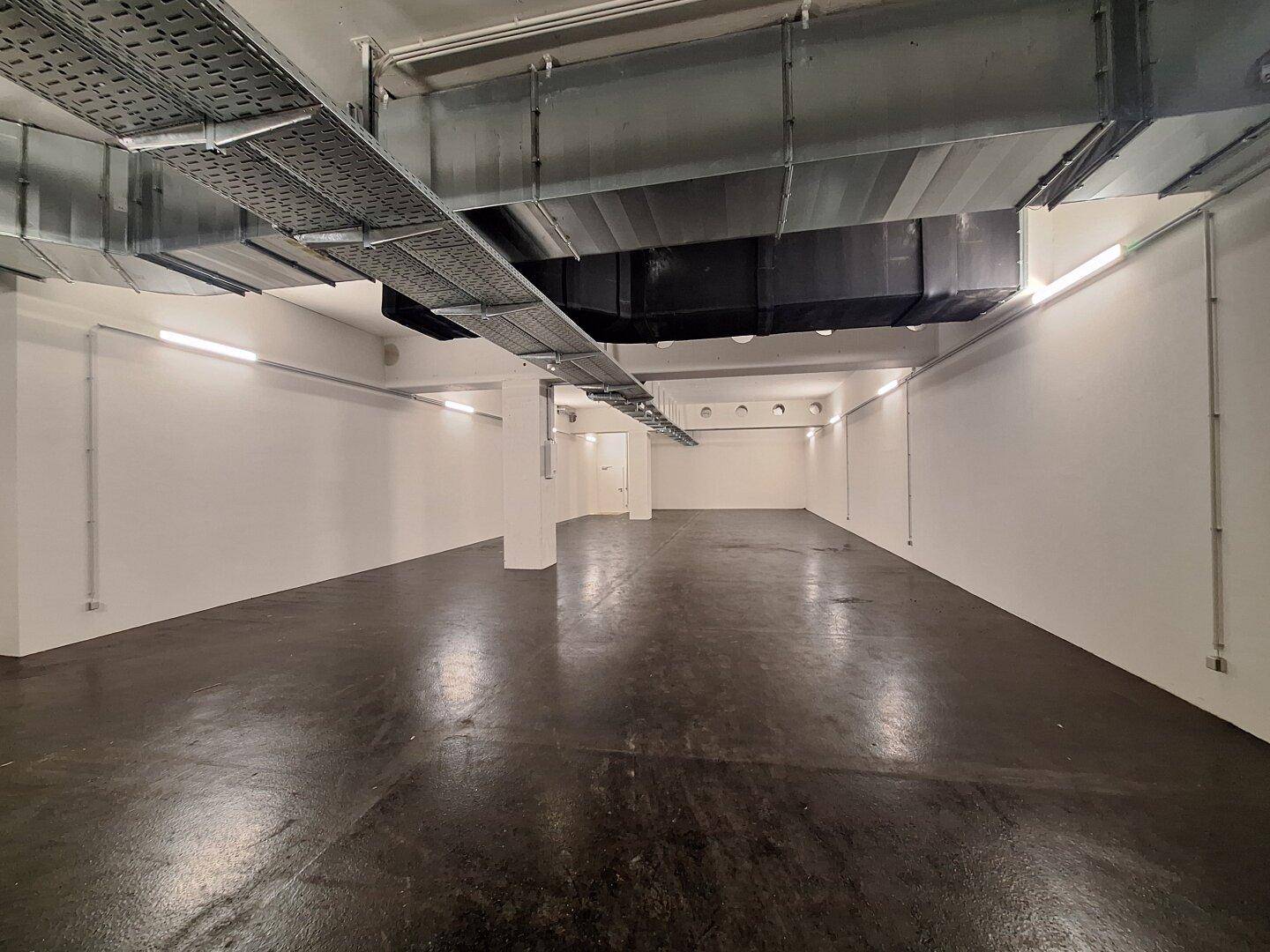 Geräumiger Lagerraum mit hohen Decken, sichtbaren Lüftungsrohren und industrieller Beleuchtung.