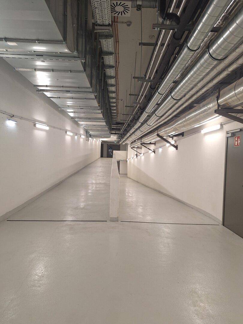 Langer, heller Kellerflur mit sichtbaren Rohren und Lüftungsanlagen an der Decke.
