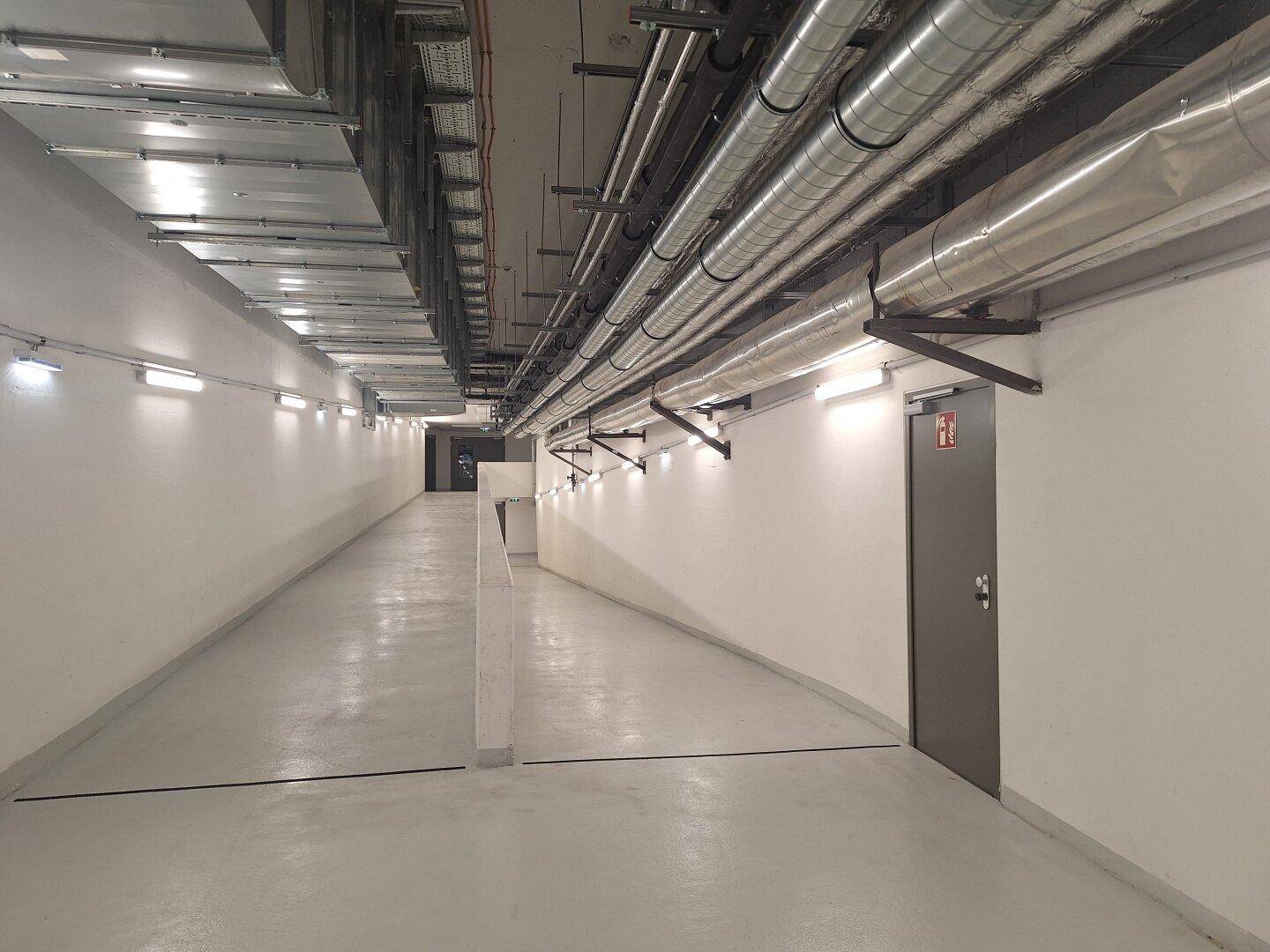 Heller Kellerflur mit sichtbaren Rohren und Lüftungsanlagen an der Decke, gut beleuchtet.