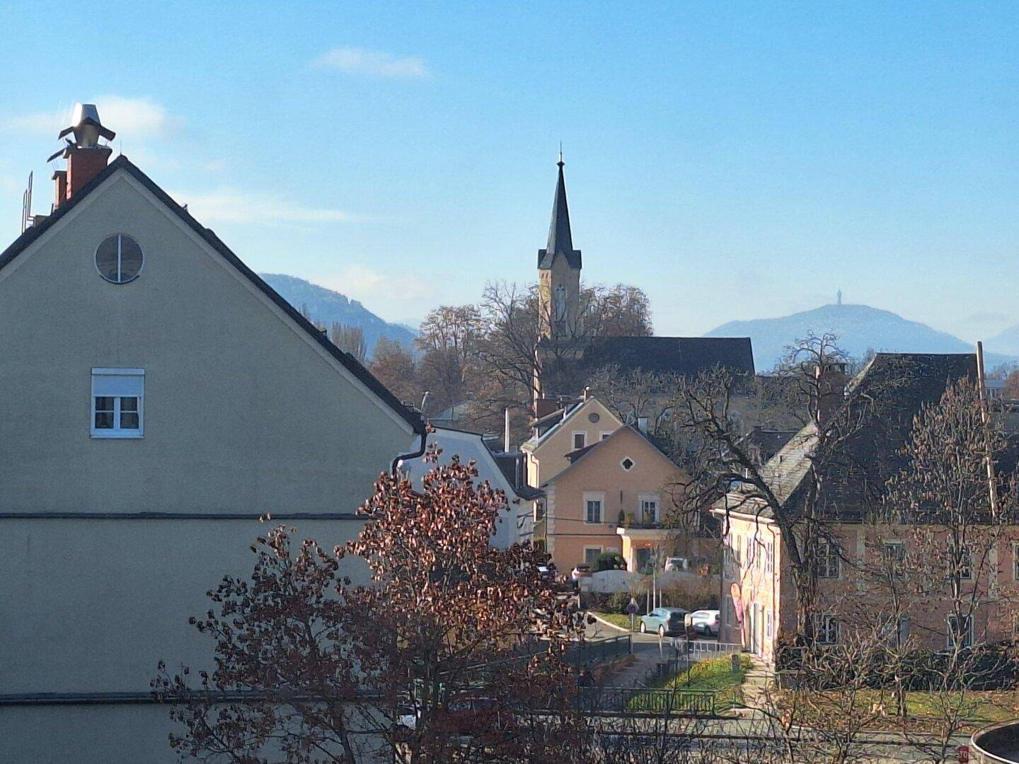 Malerischer Blick auf eine Stadt mit Kirchturm und Bergen im Hintergrund unter blauem Himmel.