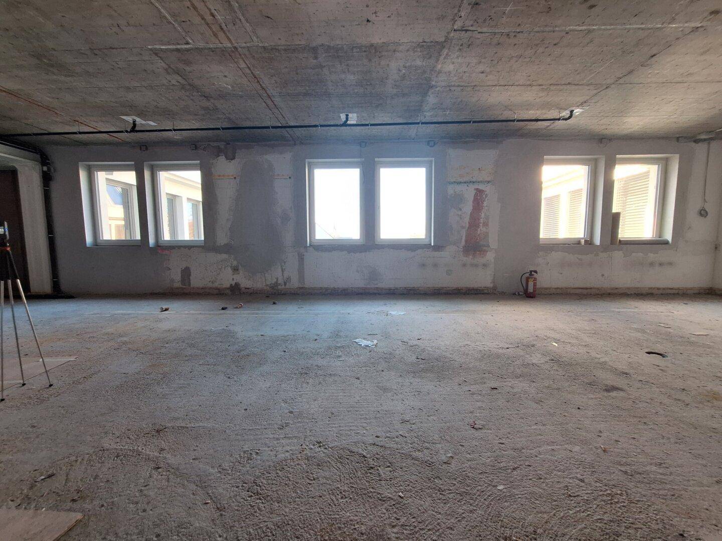 Geräumiger, unfertiger Büroraum mit Betonboden und vielen Fenstern, ideal für individuelle Gestaltung.