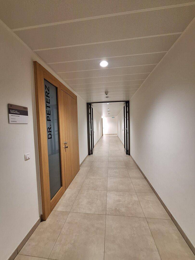 Langer, heller Flur mit beigen Fliesen und einer Holztür mit Glasausschnitt, die zu einem Büro führt.