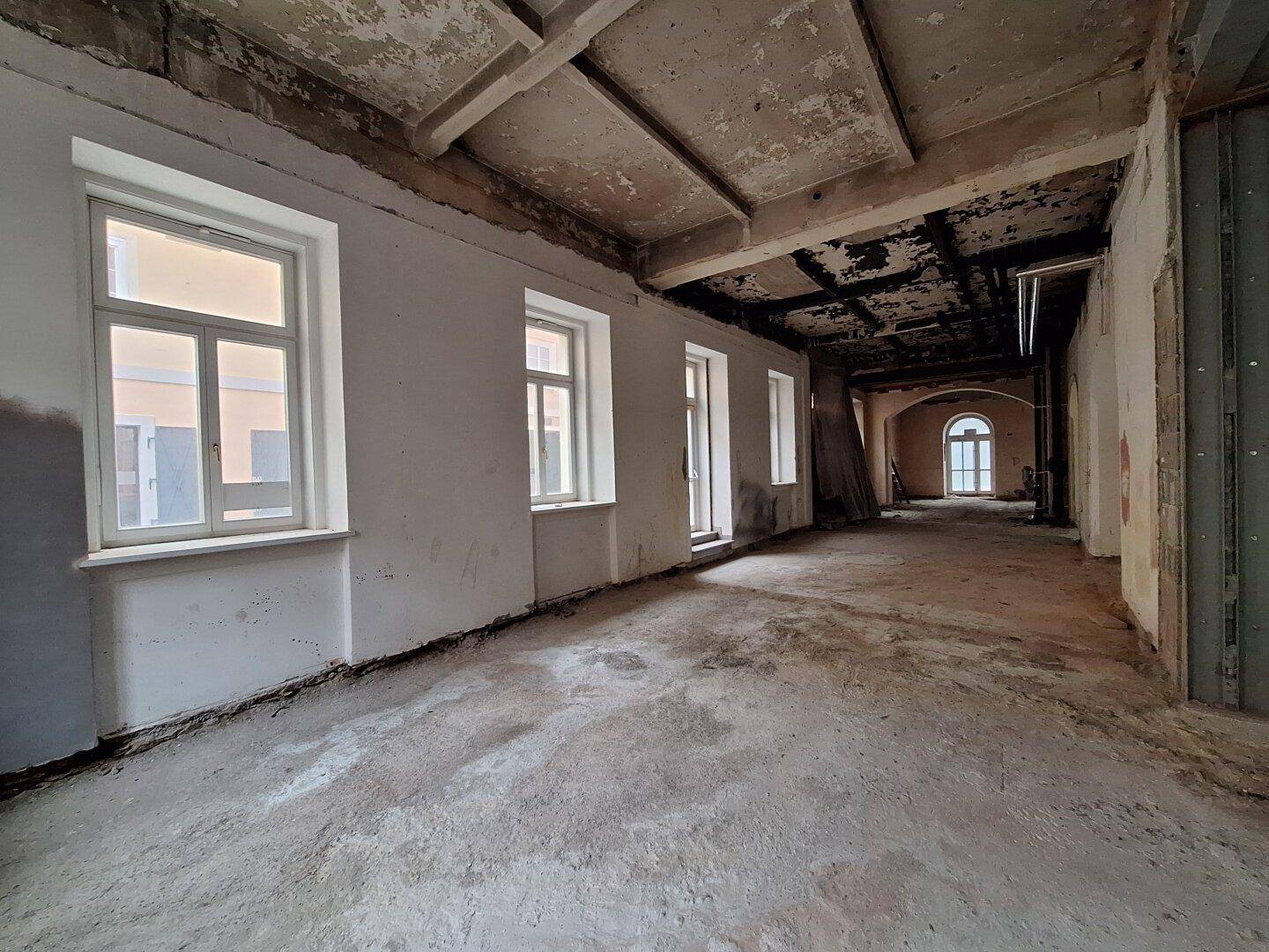 Großer, offener Raum in einem Gebäude, das sich in einem Zustand der Renovierung befindet, mit sichtbaren Fenstern und Rohbau.