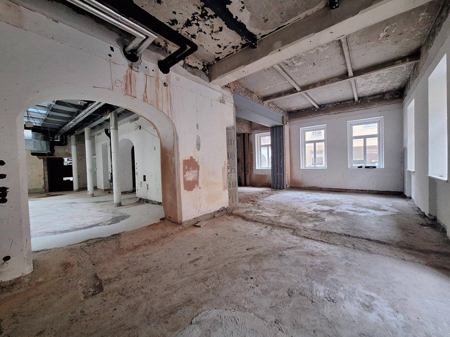 Großer, offener Raum in einem Gebäude, das sich in einem Zustand der Renovierung befindet, mit sichtbaren Fenstern und Rohbau.
