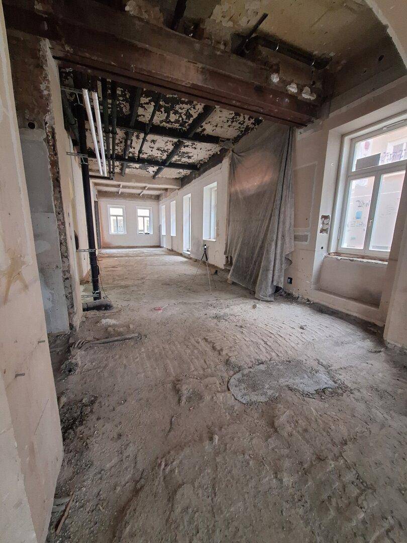 Langer, offener Raum in einem Gebäude, das sich in einem Zustand der Renovierung befindet, mit mehreren Fenstern und sichtbaren Leitungen.