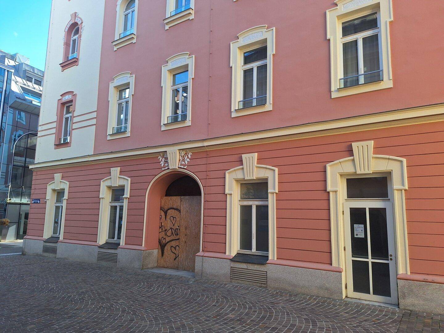 Seitenansicht eines Gebäudes mit rosa Fassade, mehreren Fenstern und einem zugenagelten Eingangsbereich.