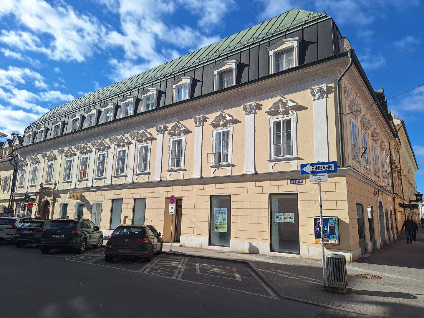 Historisches Gebäude mit Geschäften im Erdgeschoss und parkenden Autos auf der Straße unter blauem Himmel.