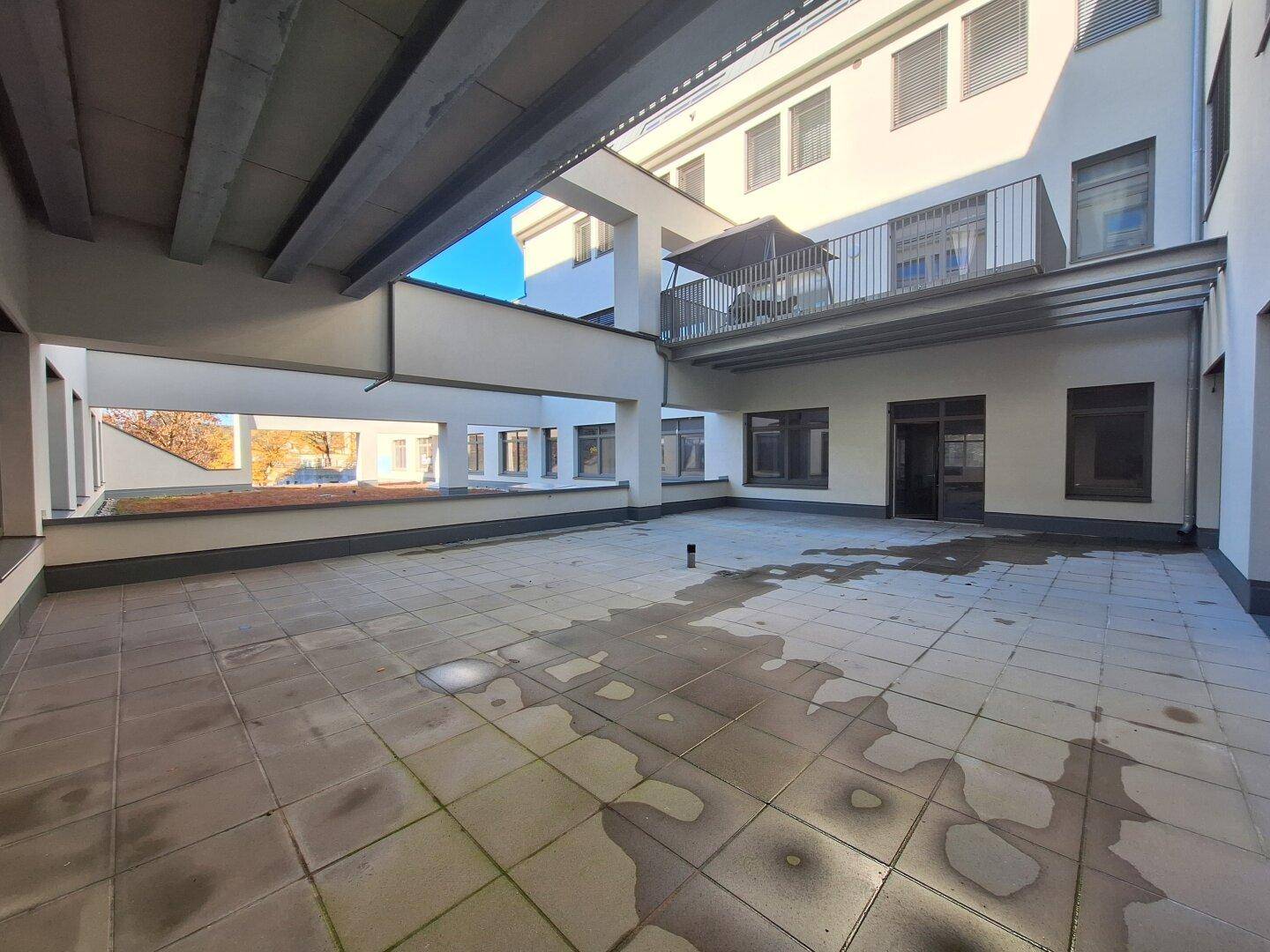 Große, gepflasterte Terrasse im Freien mit Blick auf die moderne Gebäudestruktur und eine verbindende Brücke.